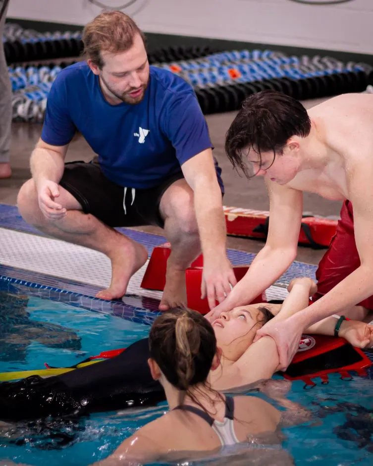 An instructor provides advice as young lifeguard trainees practice using safety equipment to remove a swimmer from a pool.
