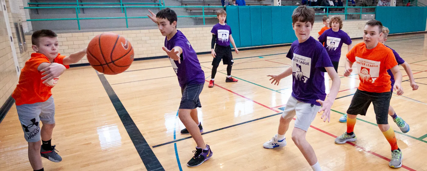 School-aged boys play basketball on an indoor court. Teams are designated with purple or orange shirts. An orange player on the left is tossing the basketball to a player out of frame as the nearest purple player with outstreched hands misses an attempt to block.