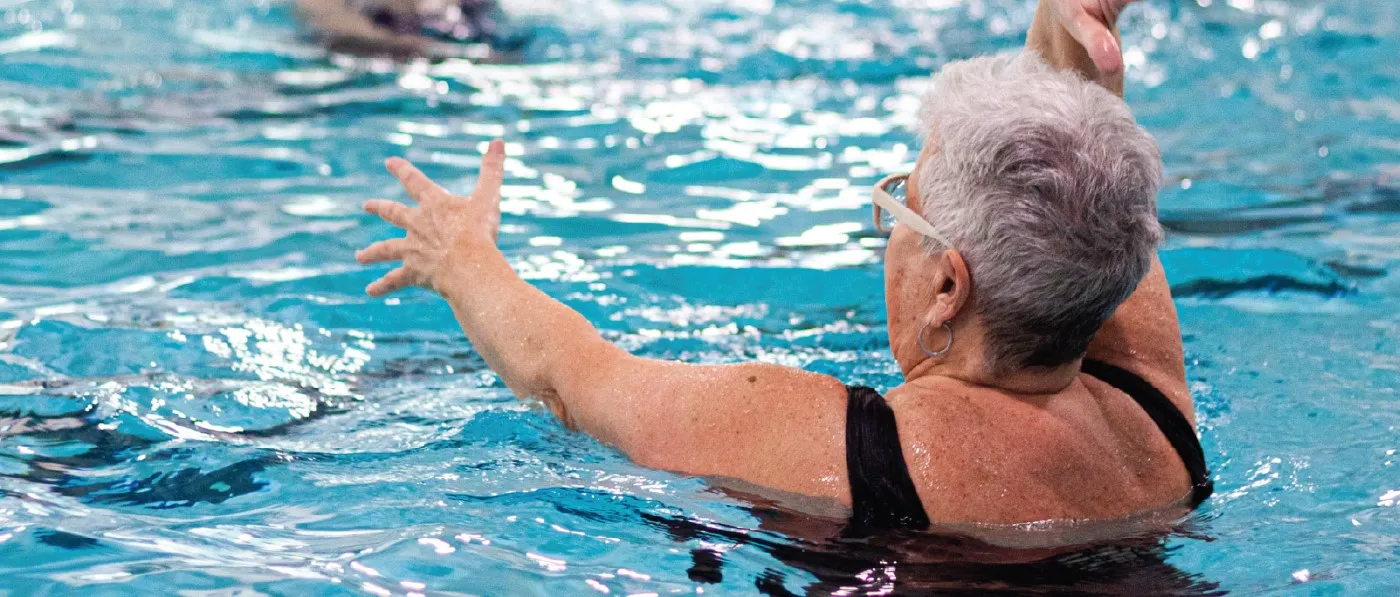 An elderly woman performs Aqua Aerobics as part of a group, moving her arms above her head while treading water as exercise.