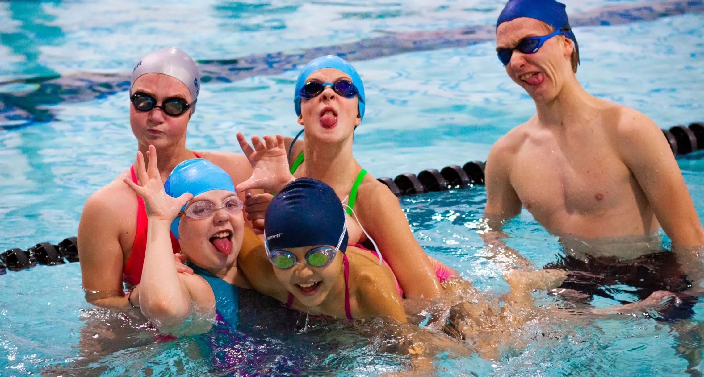 Five older children or young teens wearing swim caps and goggles make goofy faces at the camera while taking a break from swimming laps.