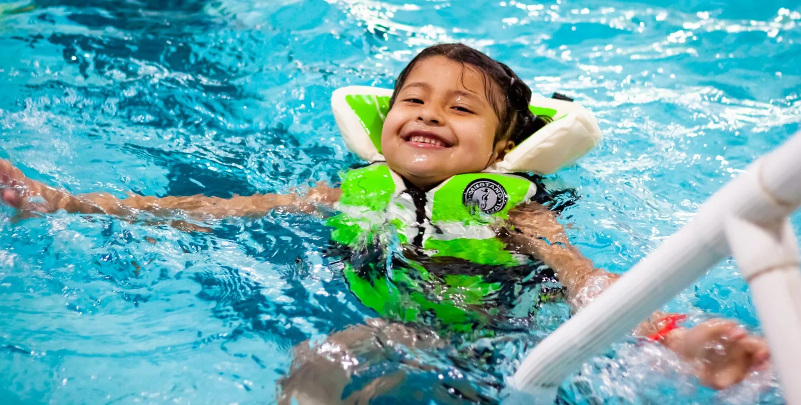 Young child smiles at camera while paddling around pool in a bulky green lifejacket.
