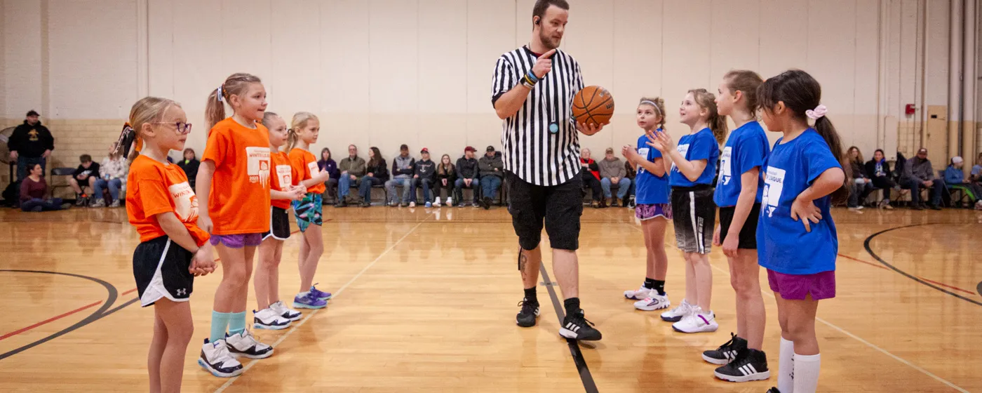 Two teams of four elementary-aged girls line up and face each other at the center of an indoor basketball court. The team on the left wears orange shirts, the team on the left wears blue shirts. Inbetween them walks an adult male referee in a black-and-white striped referee shirt, holding a basketball and speaking. Out of focus along the far wall sits a line of families who are attending the game.