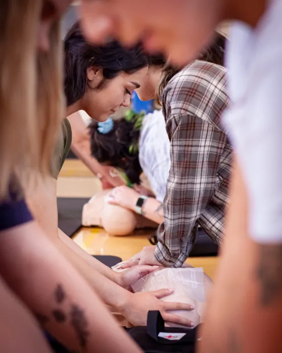 Out-of-focus bodies frame a young woman preparing to practice CPR on a dummy. She is practicing covering the fake mouth with a clear plastic guard as her training partner performs chest compressions.