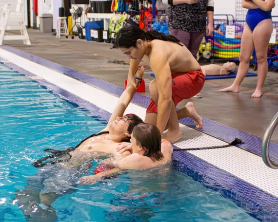 A man pretends to be unconcious in a pool during a lifeguard training exercise. A lifeguard is in the pool keeping him afloat while another lifeguard kneels at the edge of a pool to grab his arm, preparing to safely lift him out.