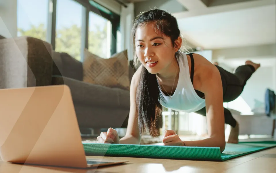 Woman on yoga mat in a home living room, performing a pose while watching YMCA360 content on her laptop.