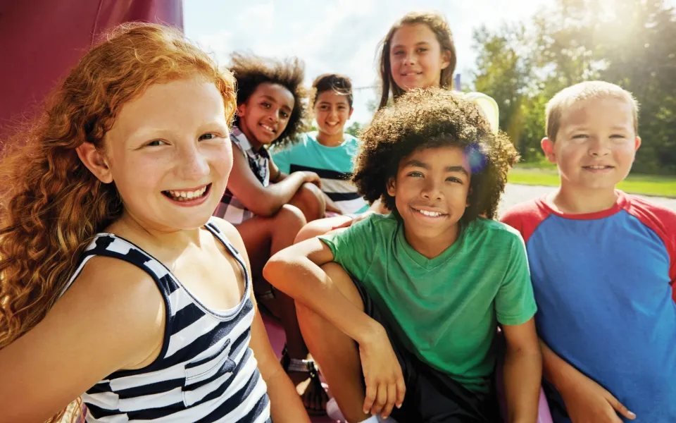 Six smiling girls and boys of middle-school age sitting outside in a sunny park, facing the camera.