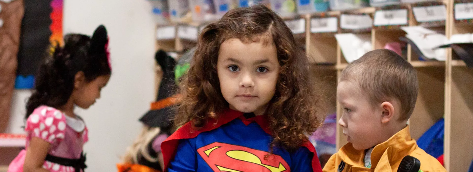 Preschoolers busy in a classroom while dressed in costume. Looking at the camera is a girl in a superhero outfit. Moving behind her are a boy dressed as a firefighter, a girl dressed as mini mouse, and a girl dressed as a witch.