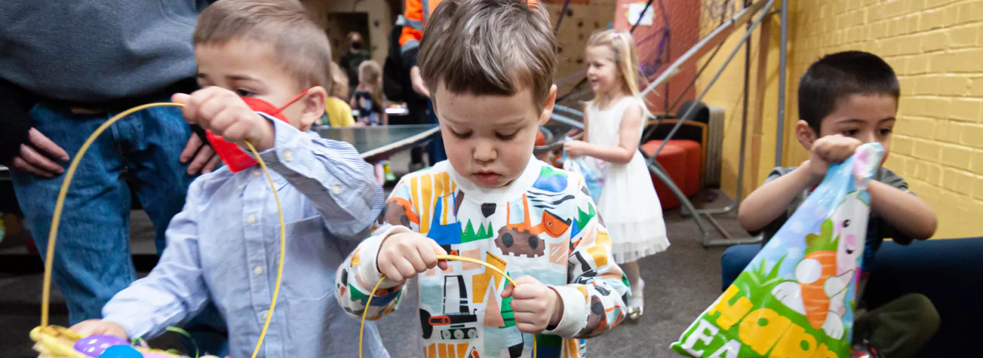 Three boys are collecting easter eggs indoors, with more children doing the same in the background. Adults are supervising. Egg baskets are full.