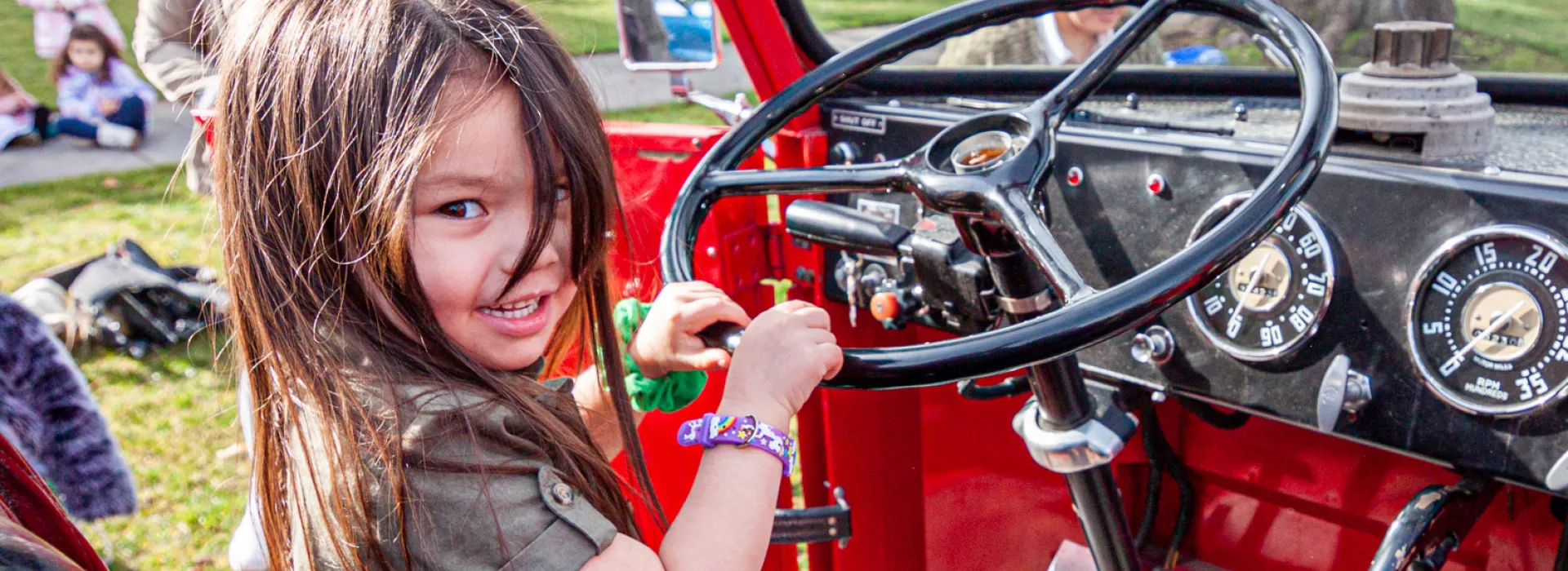 A preschool girl sits in the drivers seat and holds the steering wheel of a historic firetruck. She smiles at the camera while other kids wait their turn.
