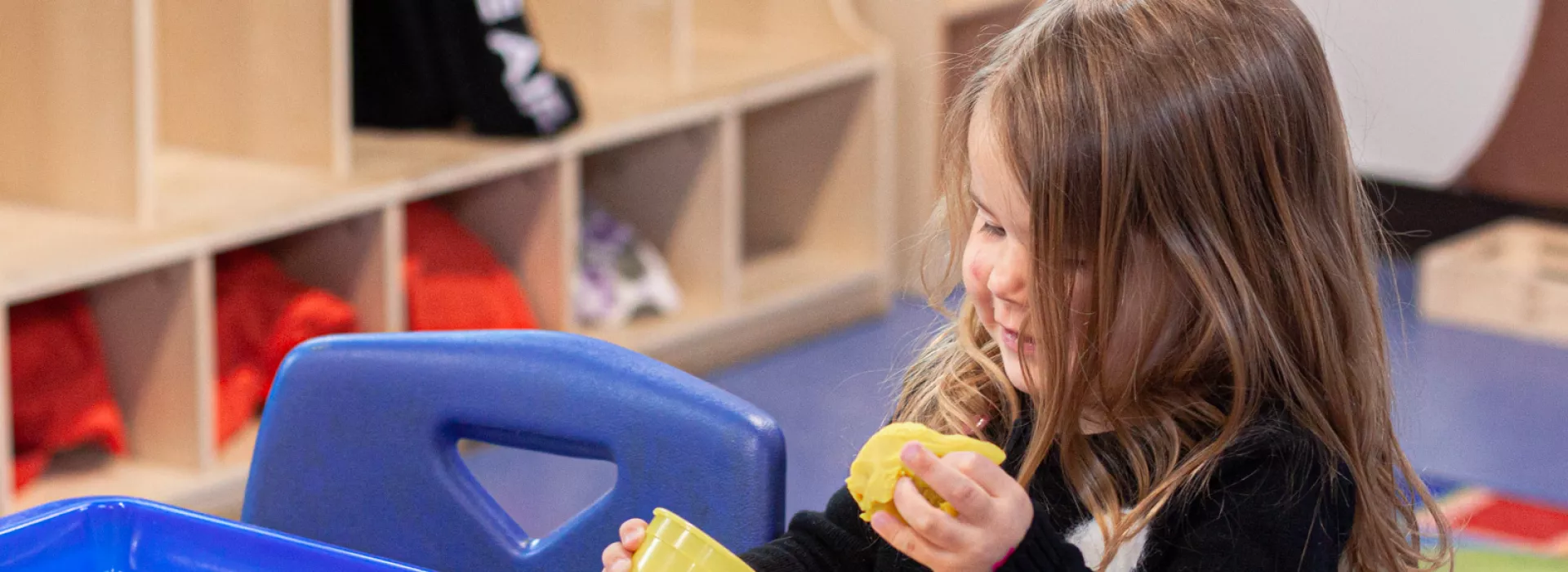 A preschool girl is playing with yellow Playdoh at a desk in a classroom. Cubbies are seen in the background behind her.