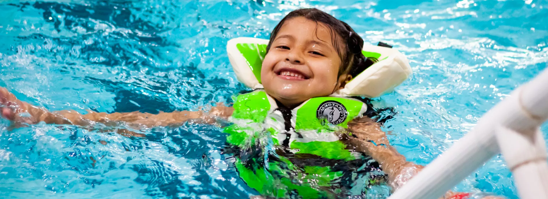 Young girl smiles at camera while floating in a blue pool. She is wearing a bright green lifejacket.