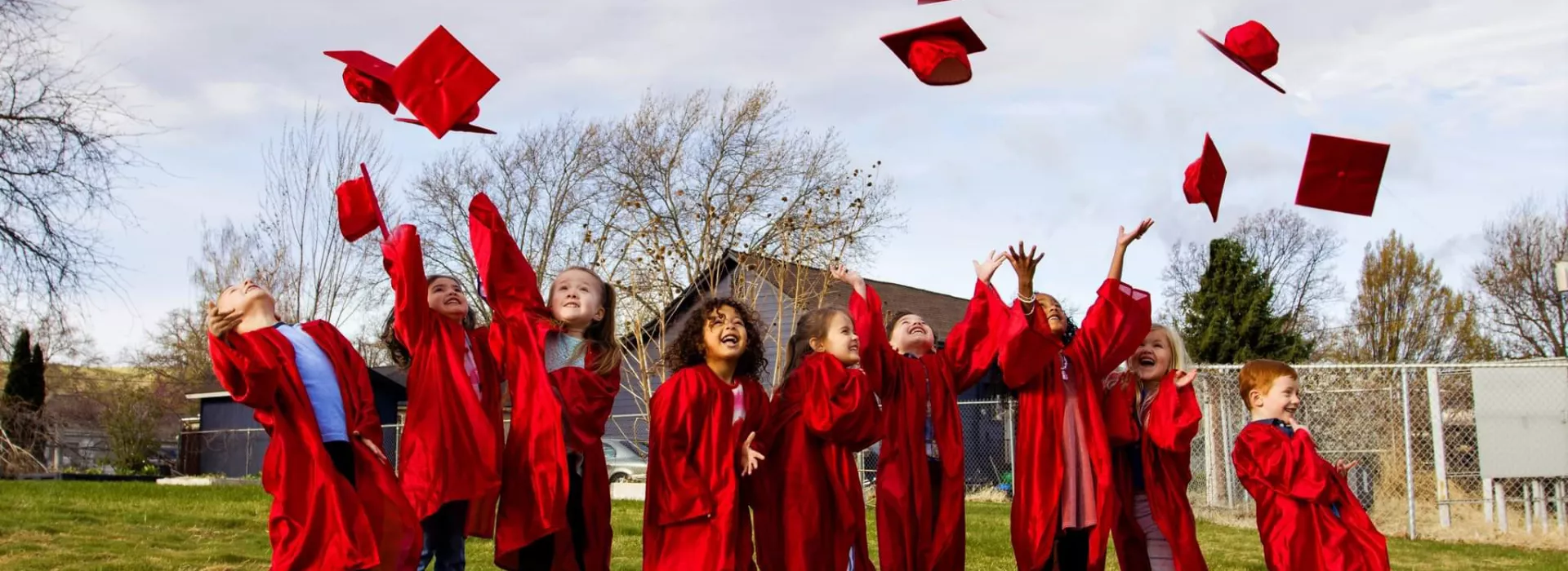 Nine preschool children standing in a line on a grassy field, wearing red gowns and joyfully tossing red graduation caps above them.