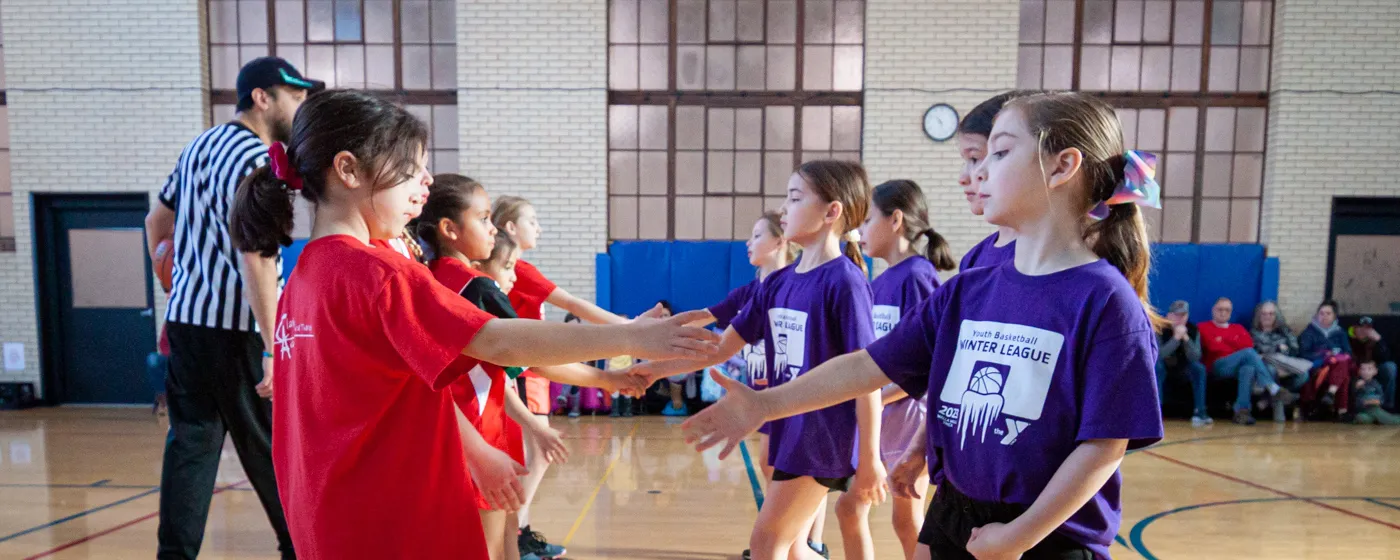 Two teams of five young girl basketball players are lined up in the center of an indoor court. The walls are off-white brick with windows that suggest the building was built in the early 1900s or earlier. The teams are approaching each other with outsteched arms to shake hands at the start of a match as their families sit along the wall to observe the game and a referee in black and white stripes supervises.