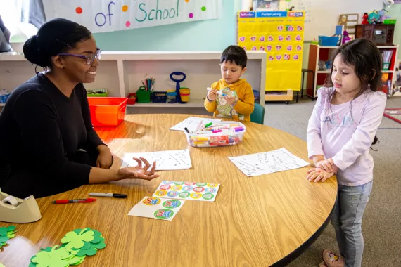A teacher sits smiling on the left behind a low, curved desk in a classroom. On the other side are two preschool-aged children interacting with paper, markers, and stickers on the desk.